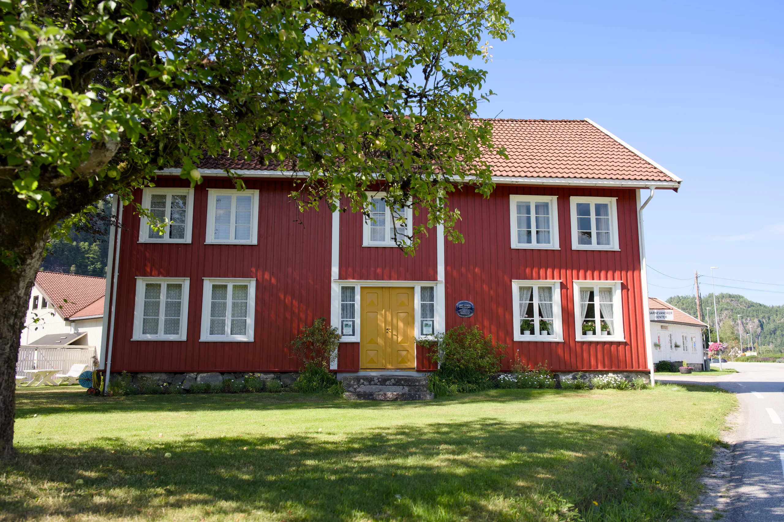 Barnevandrersenteret, et rødt toetasjes trehus med gul inngangsdør, under et stort tre en sommerdag