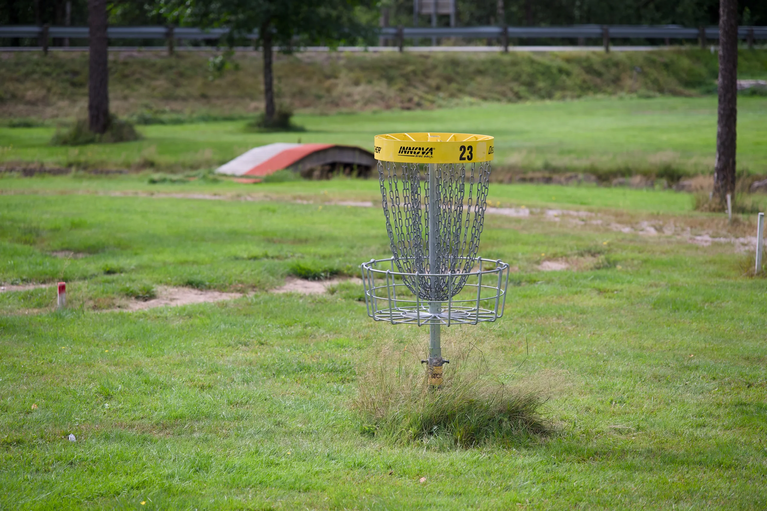 En gul frisbeegolfkurv med nummer 23 på en gressbane, med trær og en vei i bakgrunnen