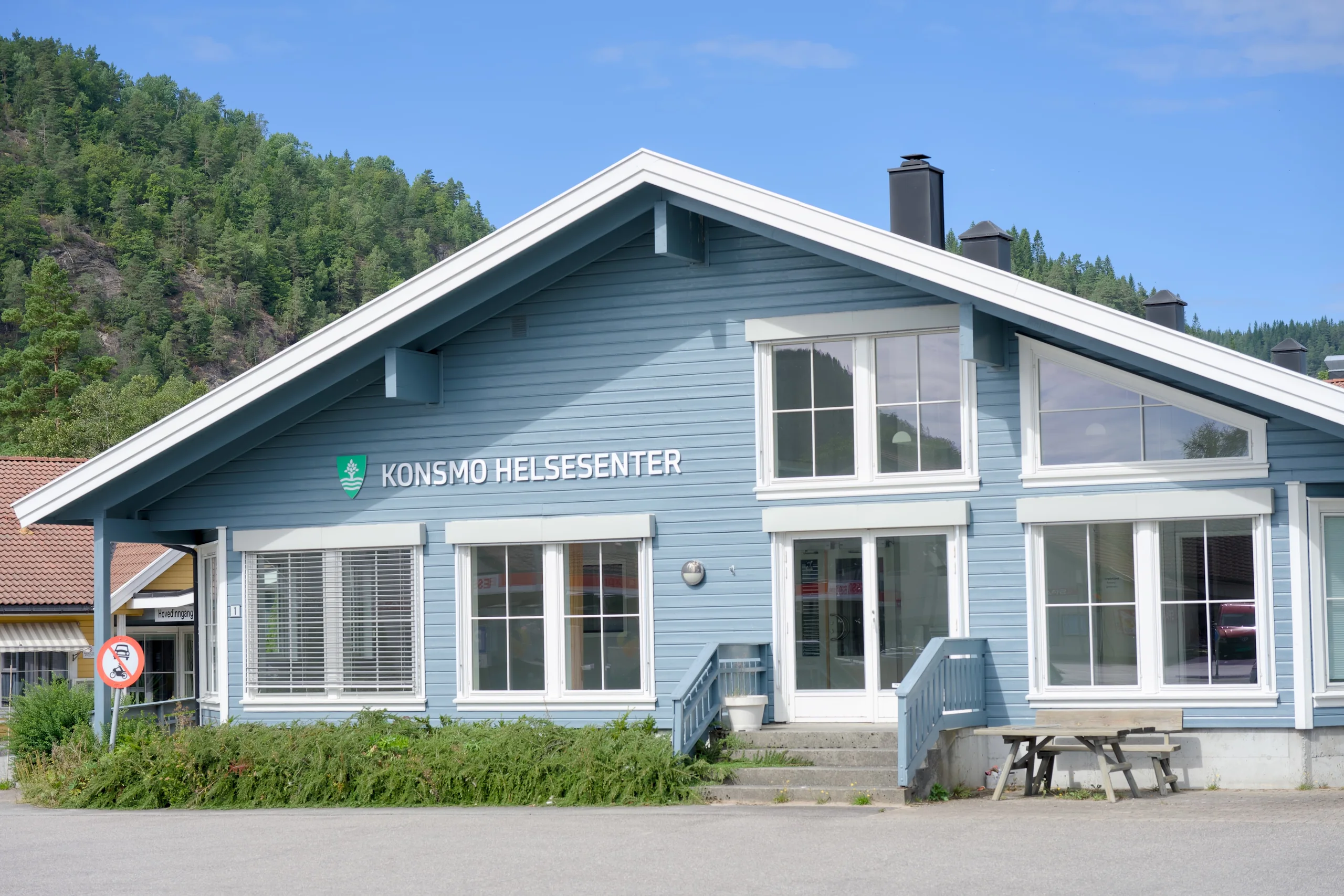 Konsmo helsesenter, et lyseblått trehus med store vinduer og en benk ved inngangen
