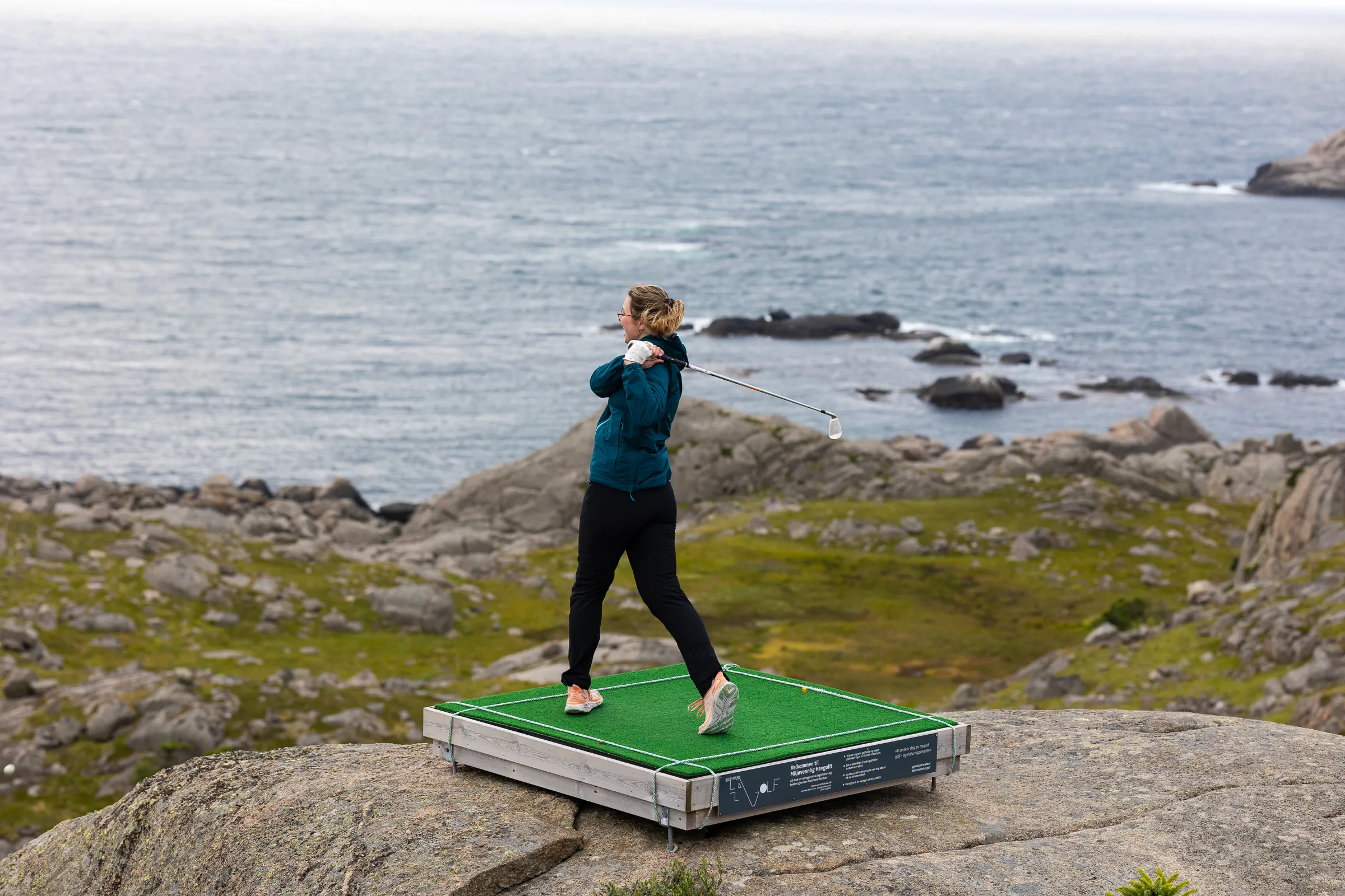 Kvinne slår ut fra en havgolf-matte på svaberget, med havet bak