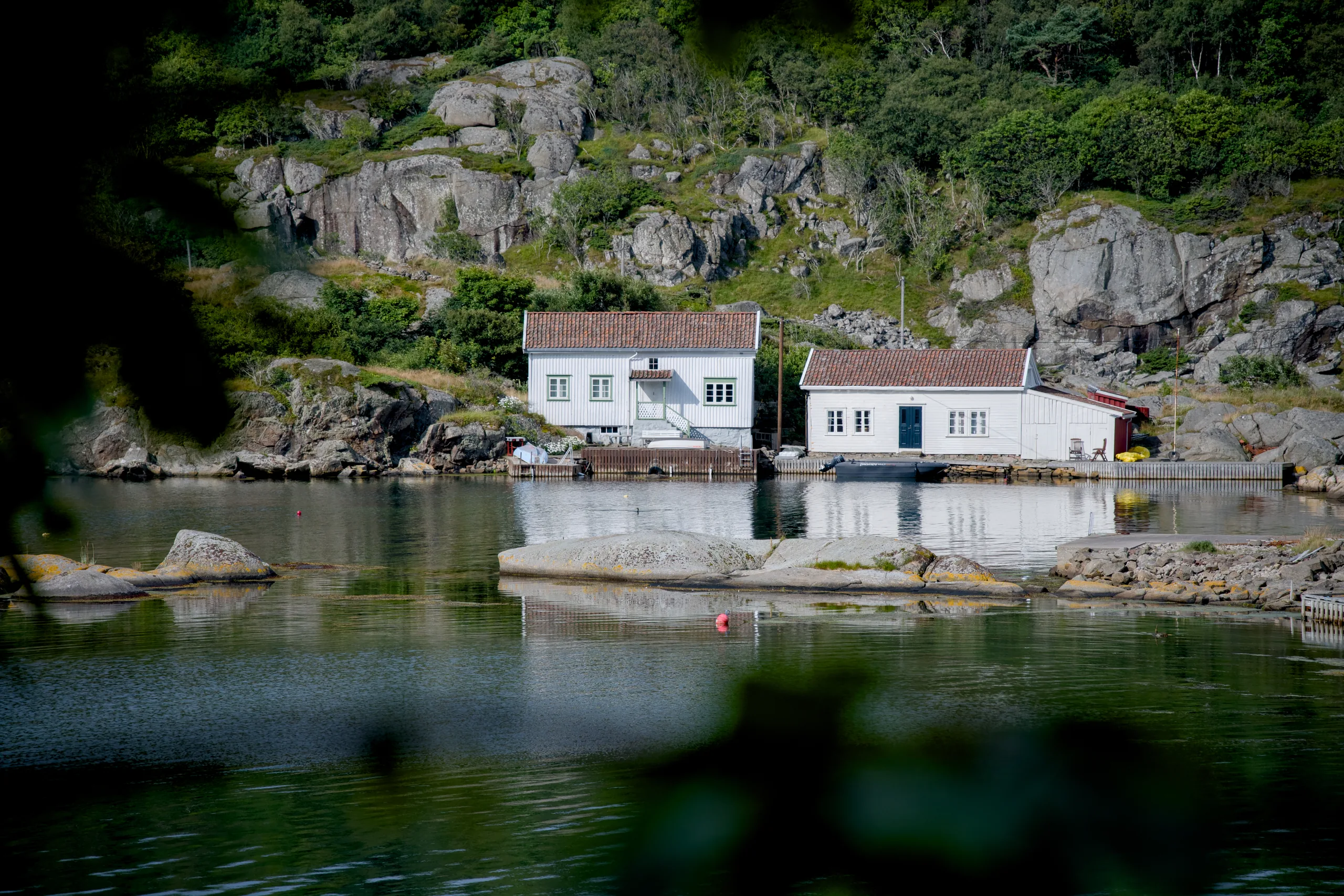 To hvite trehus med teglsteinstak ved en brygge under bratte svaberg og skog, sett over stille vann gjennom løvverk