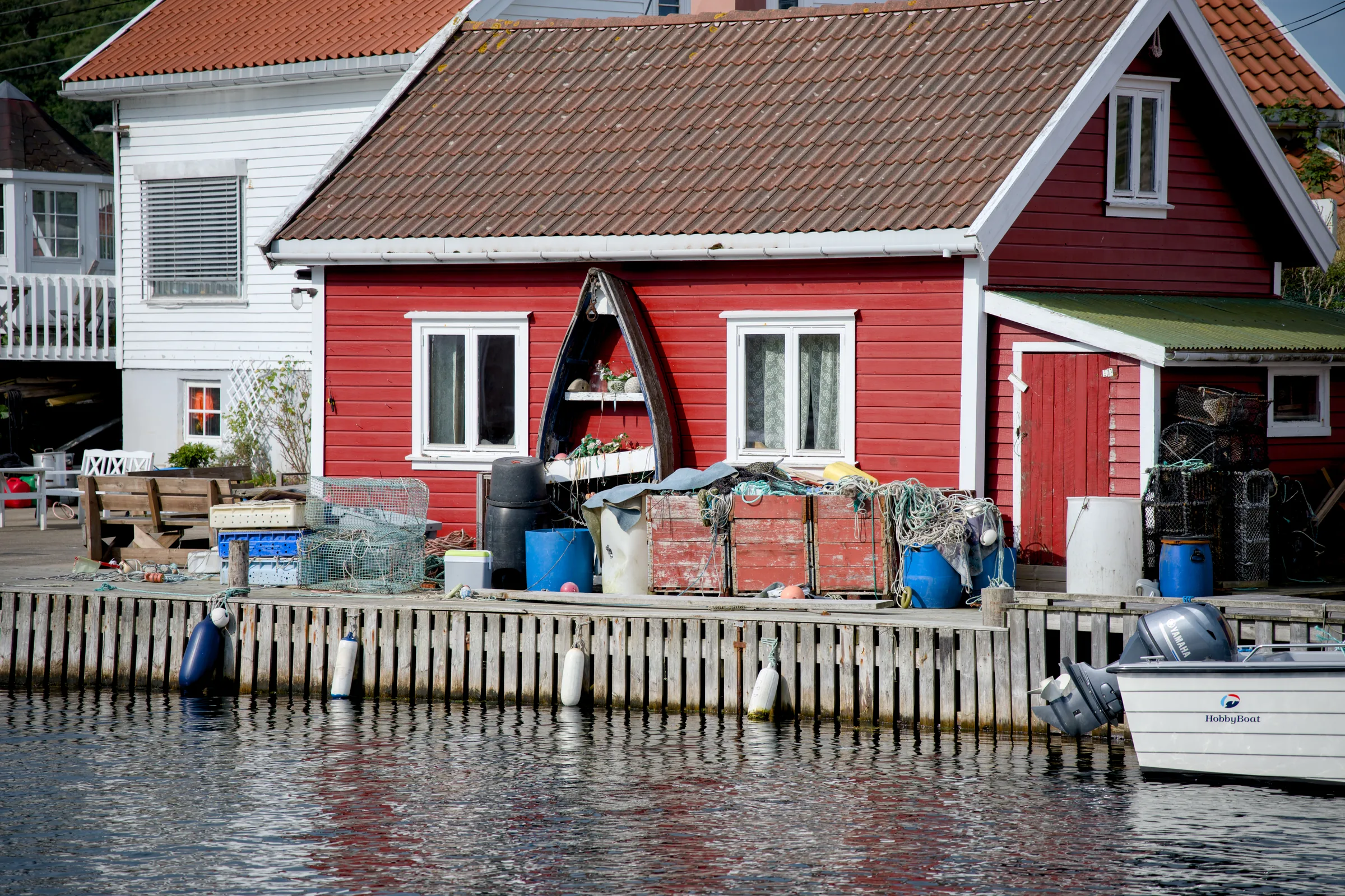Rødt naust med teglsteinstak på brygga, med fiskekasser, garn, teiner og blå tønner foran og en båt fortøyd ved kaikanten