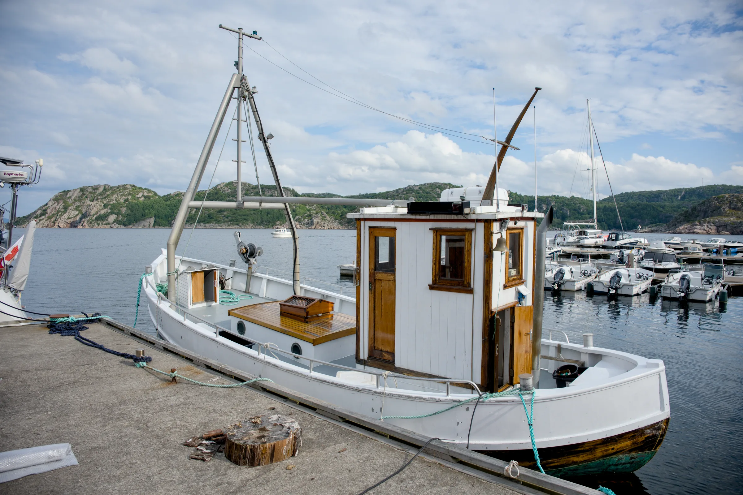 En hvit trebåt med styrehus fortøyd ved kaia, med småbåthavna og svaberg bak
