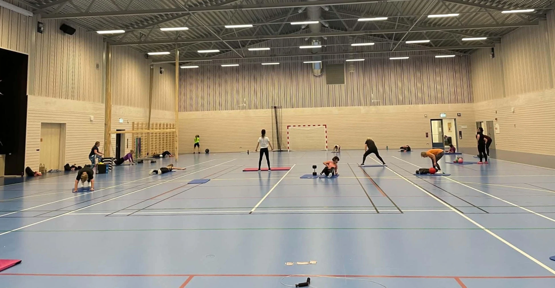 Barn og voksne trener på gulvet i en stor, lys idrettshall med merkede baner