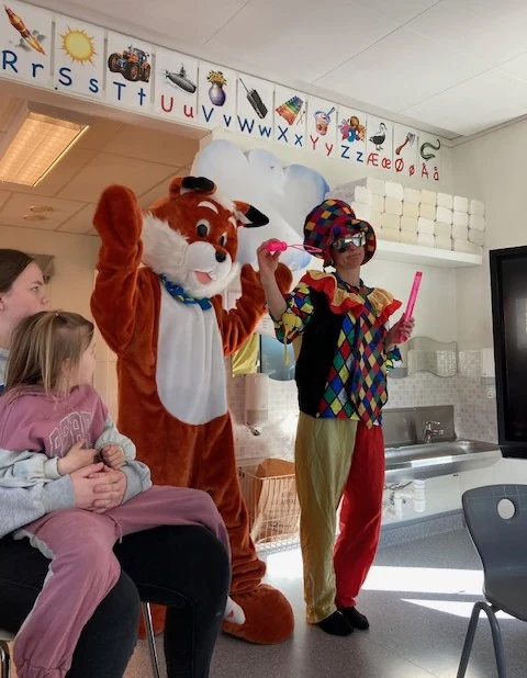 Barn og voksne i et klasserom, med en rev i maskeradedrakt, en bamse og en klovn som vinker