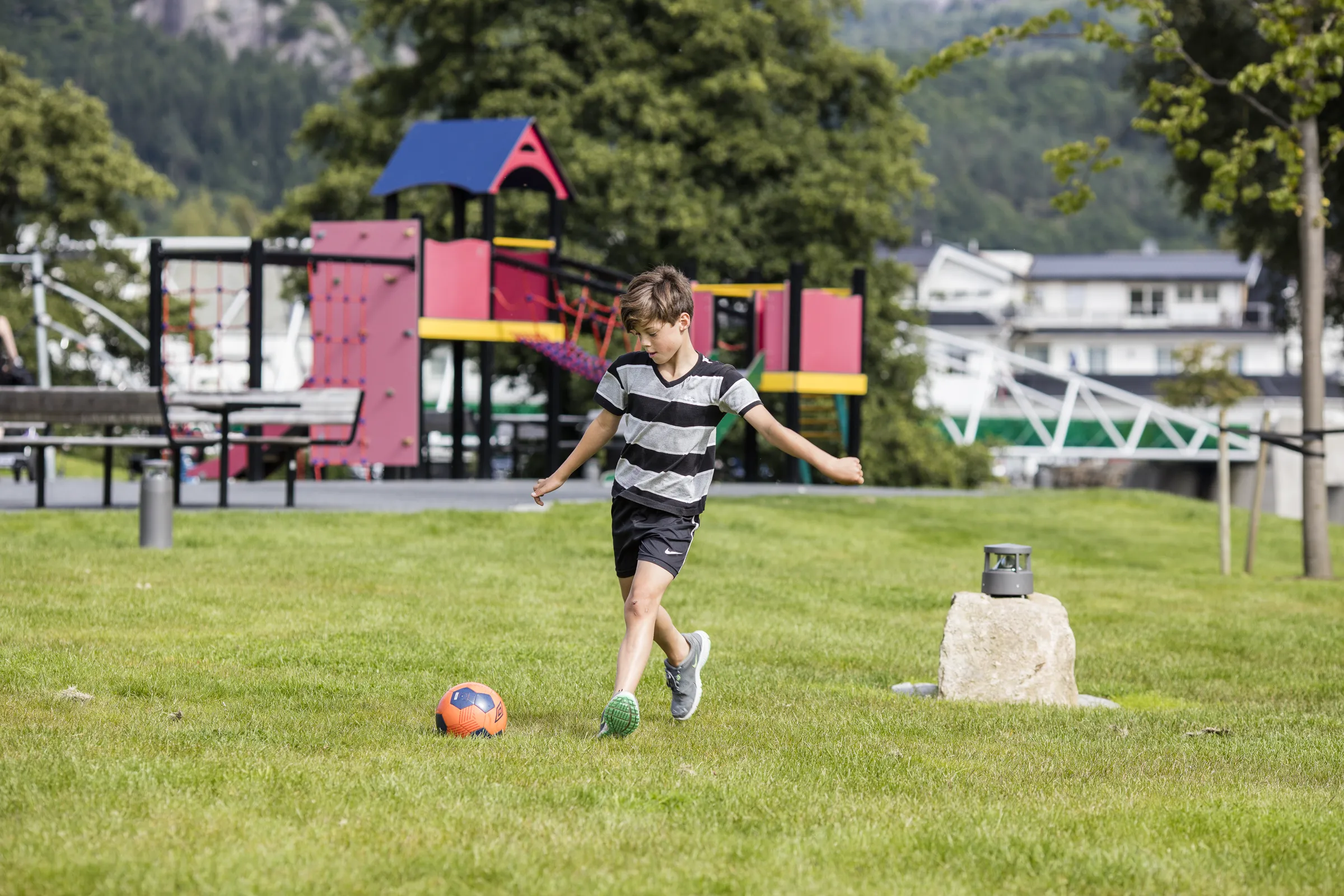 Gutt spiller fotball i Kvinaparken, med lekeapparatene bak