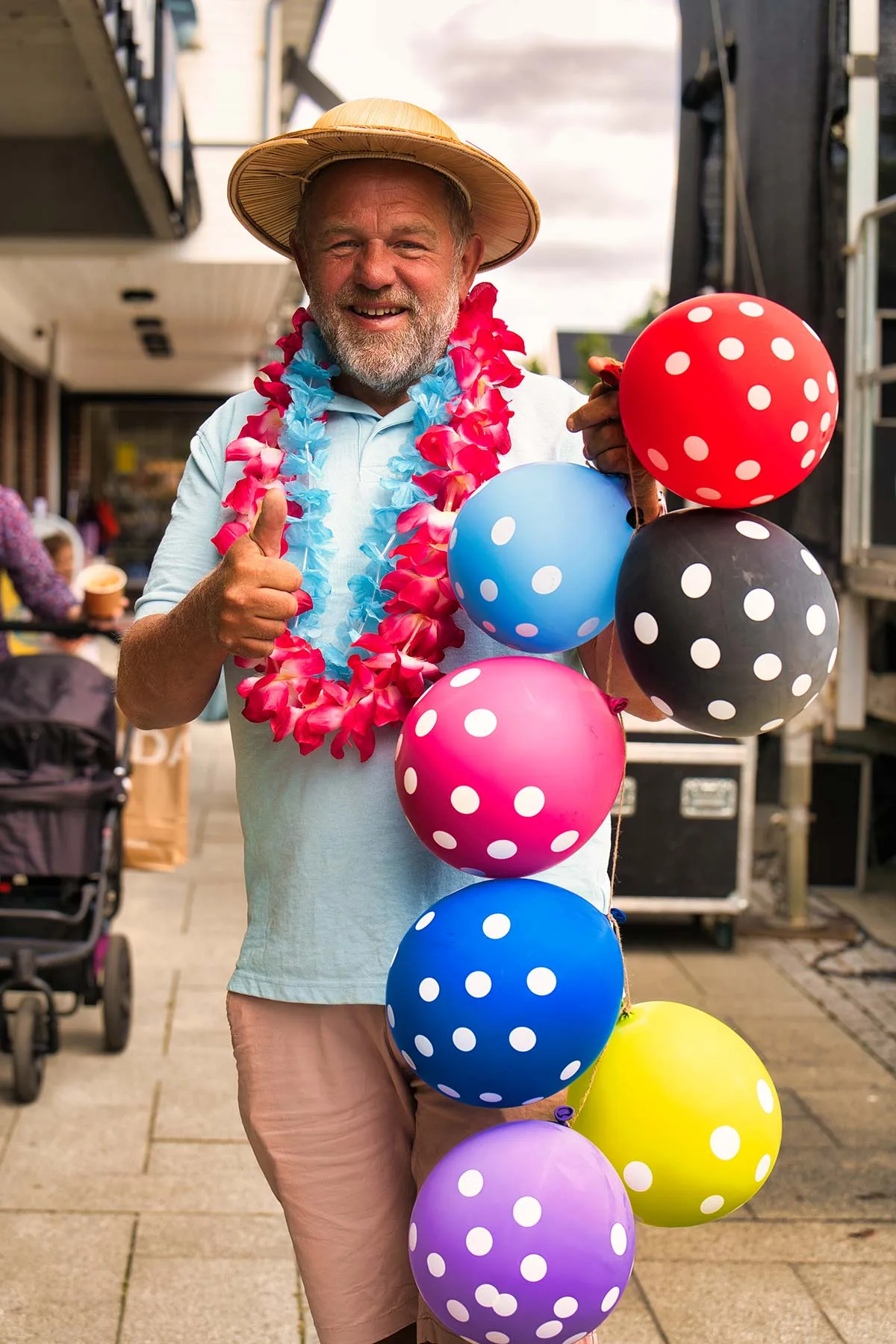 En smilende mann med stråhatt og blomsterkrans holder en klase prikkete ballonger i gågata under Byfesten