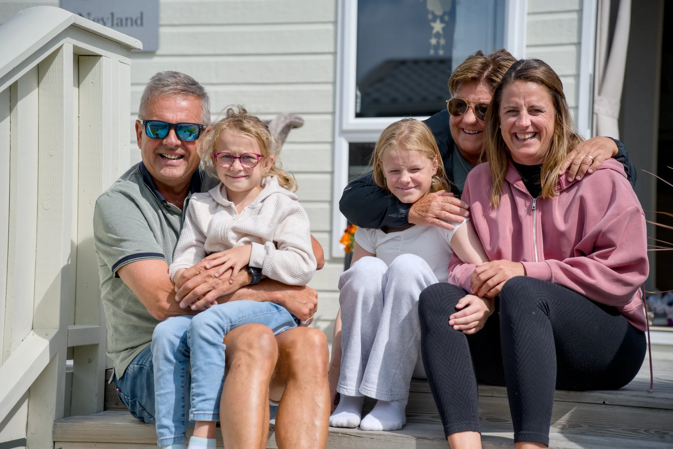 En familie med tre generasjoner sitter tett sammen på en benk foran et hvitt trehus