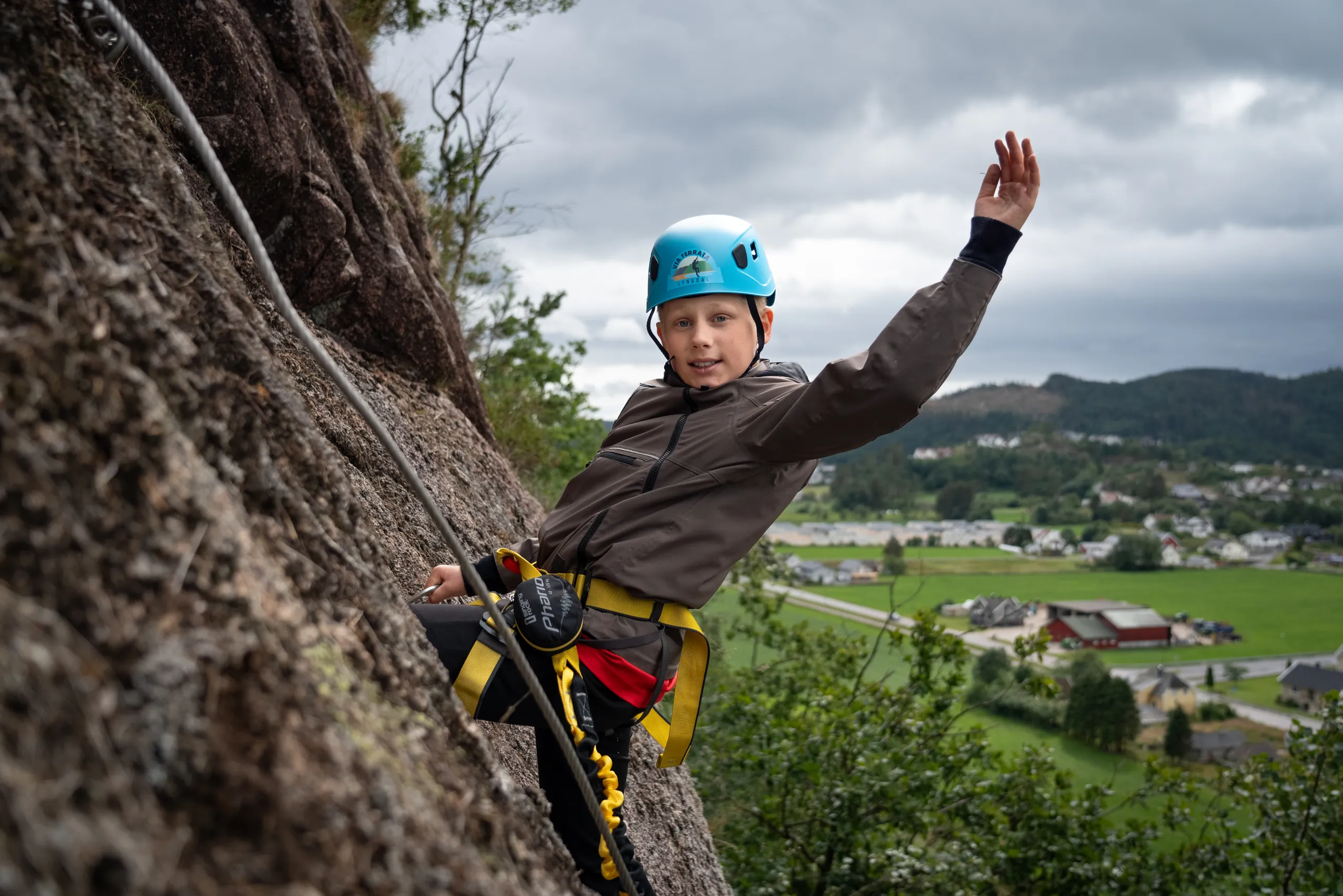 En gutt med hjelm og klatresele vinker fra fjellveggen på Via Ferrata, med Lyngdal i bakgrunnen