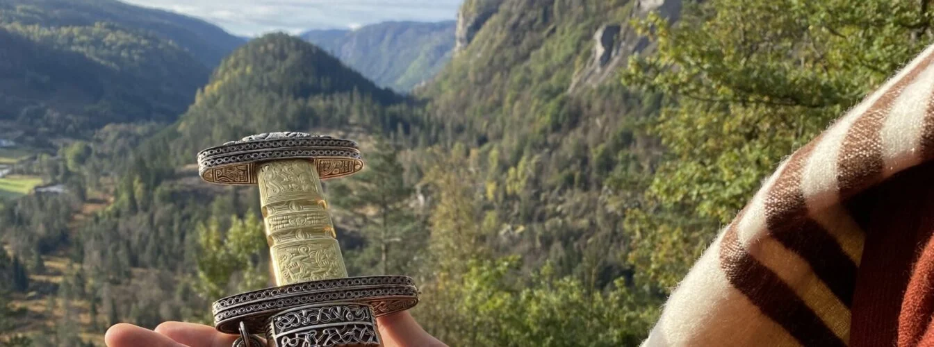 En hånd holder en kopi av håndtaket på Snartemosverdet, med dalen og skogkledde fjell i bakgrunnen