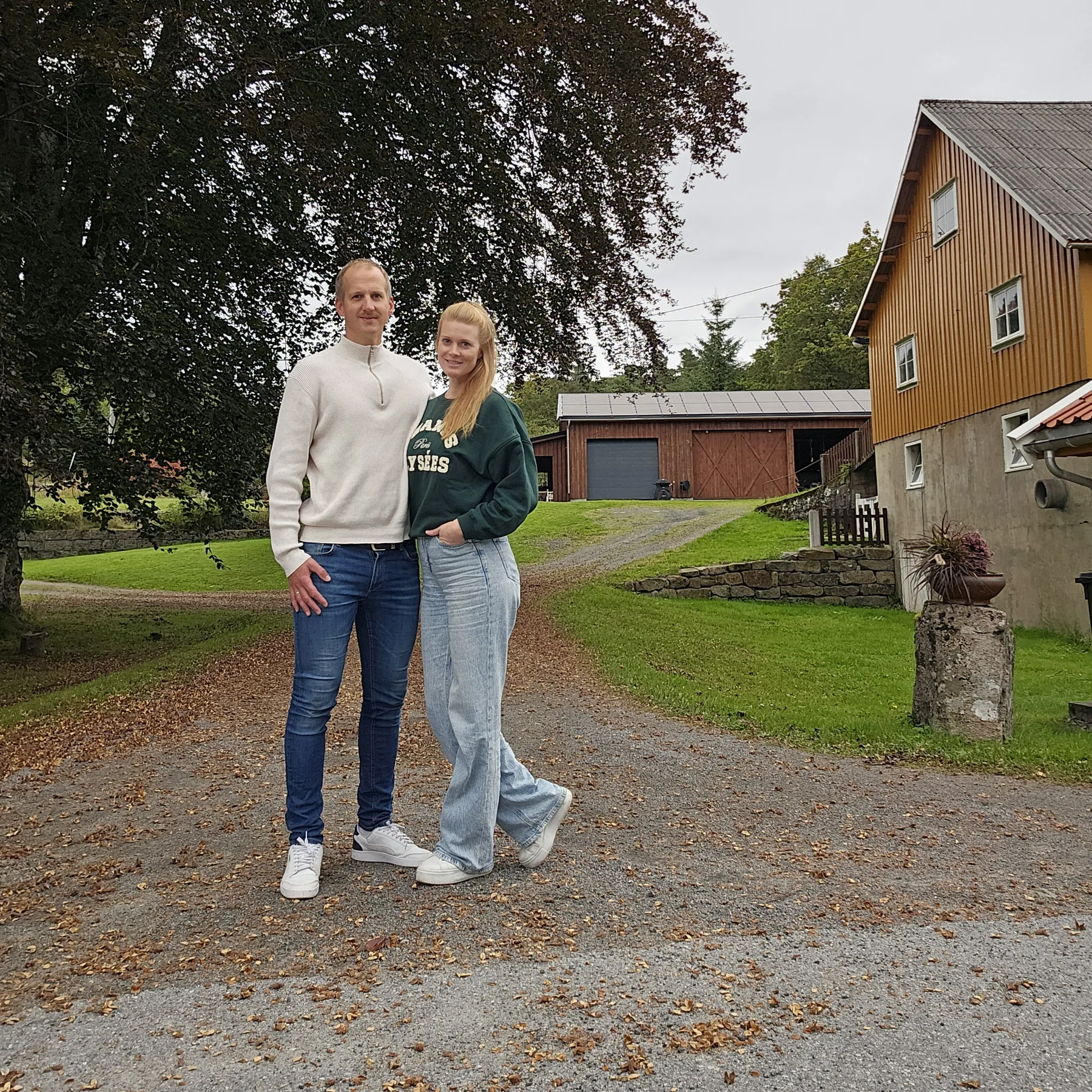 Nils Edvin Numme og kona står arm i arm på gårdsplassen, med et stort bøketre, en låve og et gult hus bak