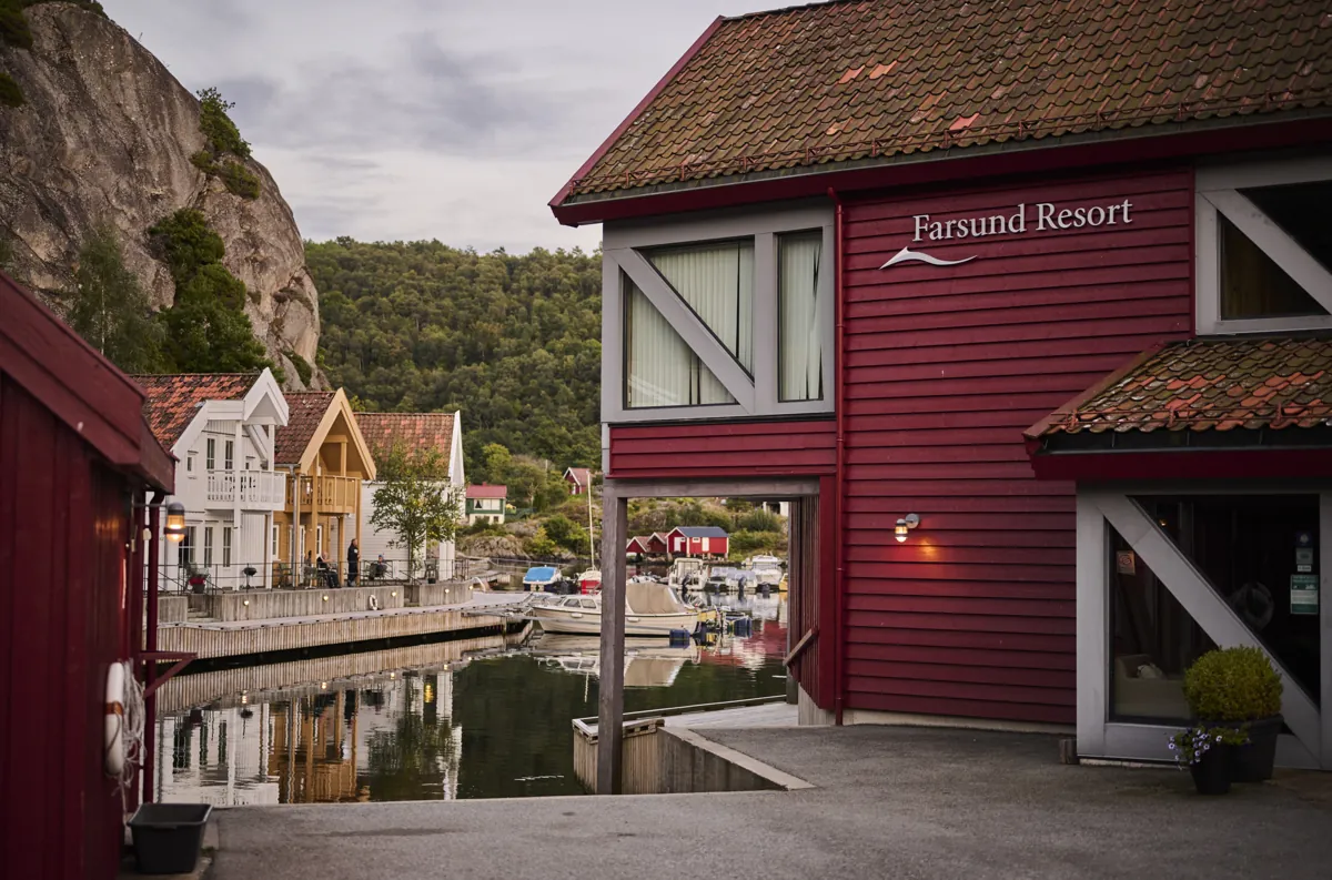 Det røde hovedbygget med skiltet Farsund Resort, med utsikt gjennom en åpning mot havna, båter og hvite hus under en bratt fjellvegg