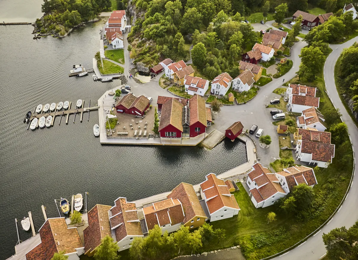 Luftfoto rett ovenfra av Farsund Resort: hvite og røde hus med teglsteinstak rundt havna, brygger med småbåter og en svingete vei