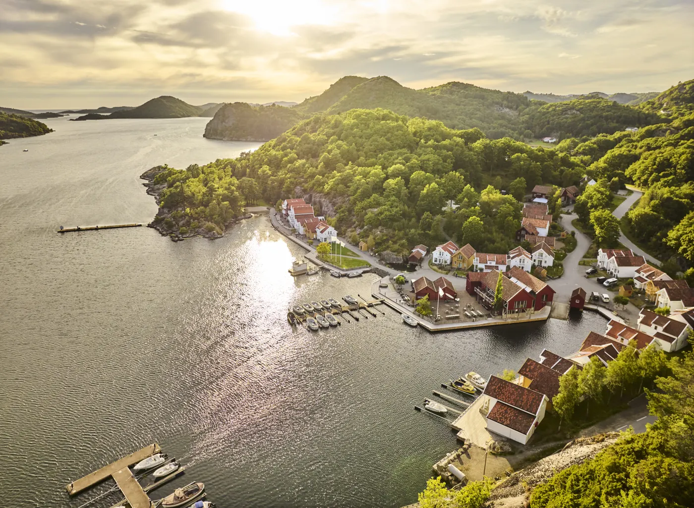 Farsund Resort fra lufta i kveldssol: røde og hvite hus rundt en liten havn med båtplasser, skogkledde åser og fjorden som åpner seg mot havet
