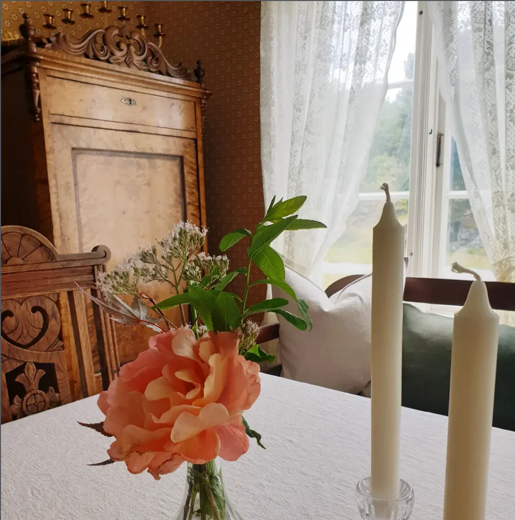 Stua i Skipperhuset: et gammelt hjørneskap i tre, blondegardiner, en rosa rose i vase og to hvite lys på et hvitt bord