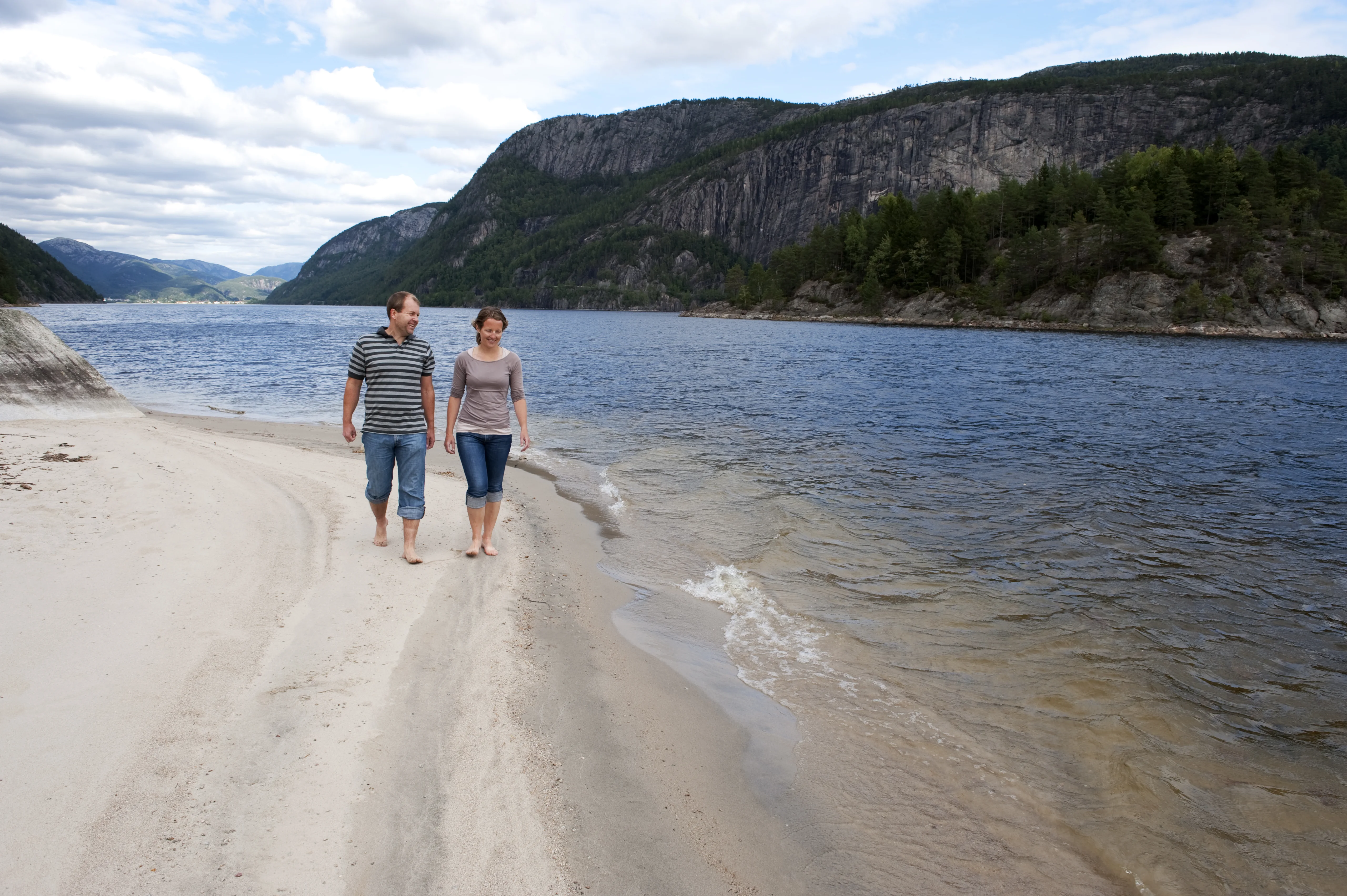 Et par går barbeint langs den hvite sandstranda ved Sirdalsvannet, med bratte fjellsider bak
