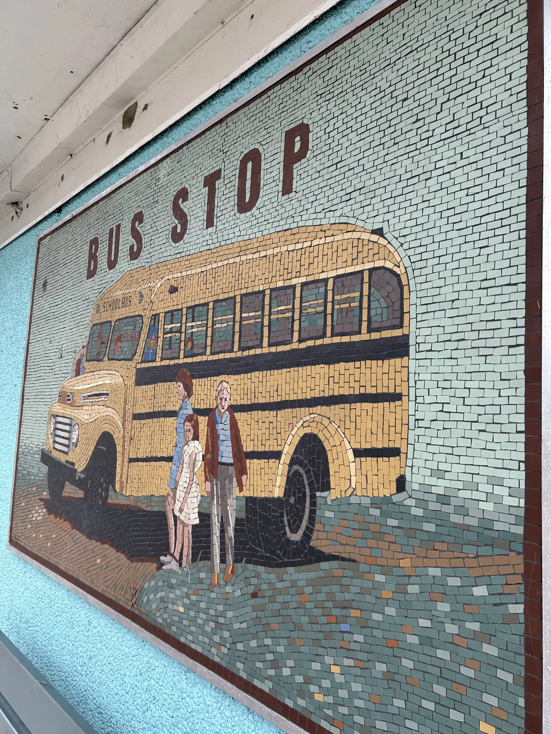 Mosaikk av en gul amerikansk skolebuss med teksten «Bus Stop» på en husvegg i Vanse