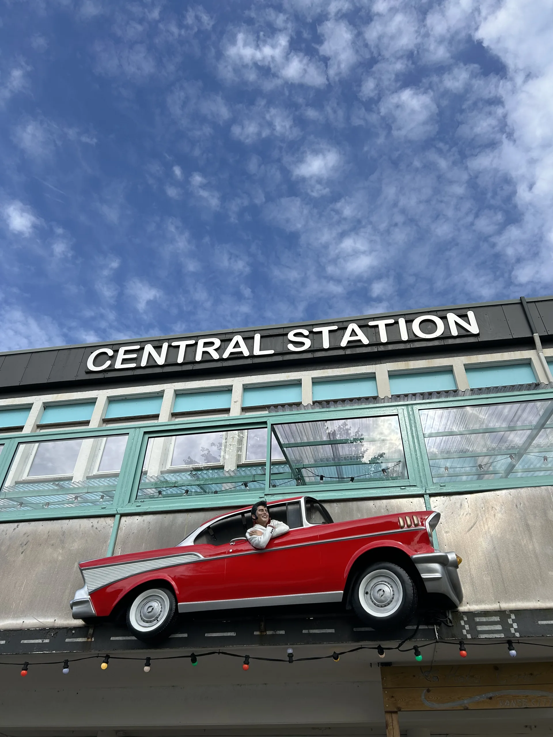 Central Station i Vanse: en rød veteranbil montert over inngangen til et turkist bygg, under blå himmel