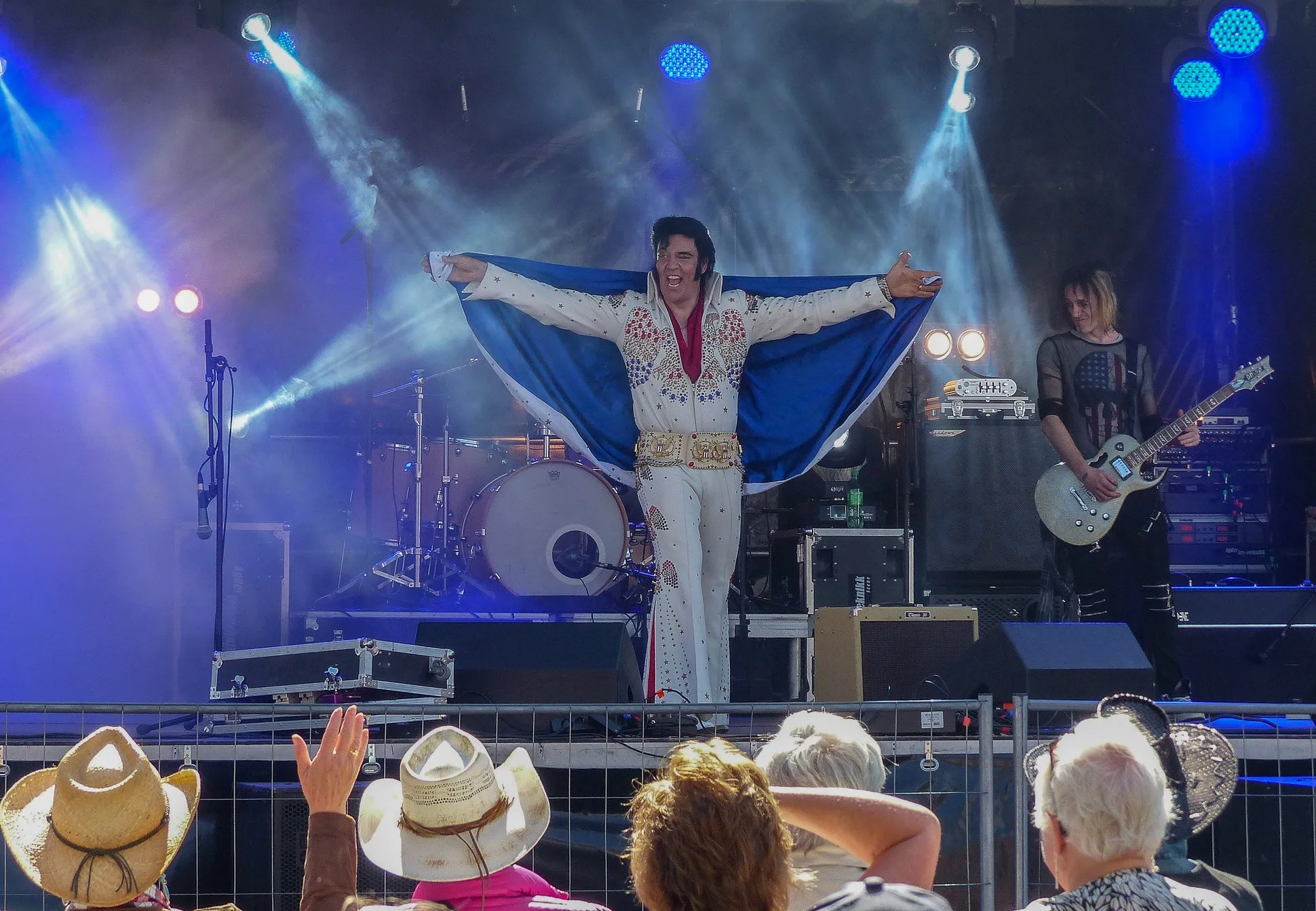 Elvis-artist i hvit dress med utstrakte armer på scenen i blått lys, med publikum i cowboyhatter foran