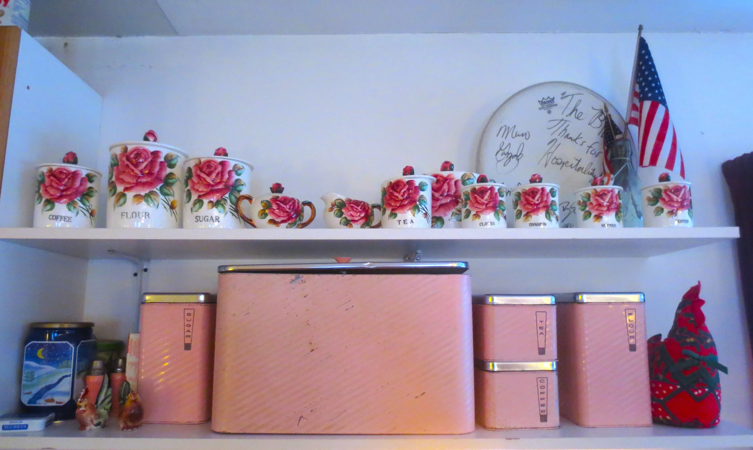 Pink retro kitchen canisters and rose-patterned mugs on shelves in the American museum apartment, with a small American flag