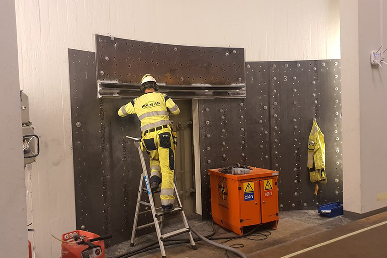 Ansatt i gul vernedress og hjelm jobber på en stige med en stålkonstruksjon inne i et anlegg