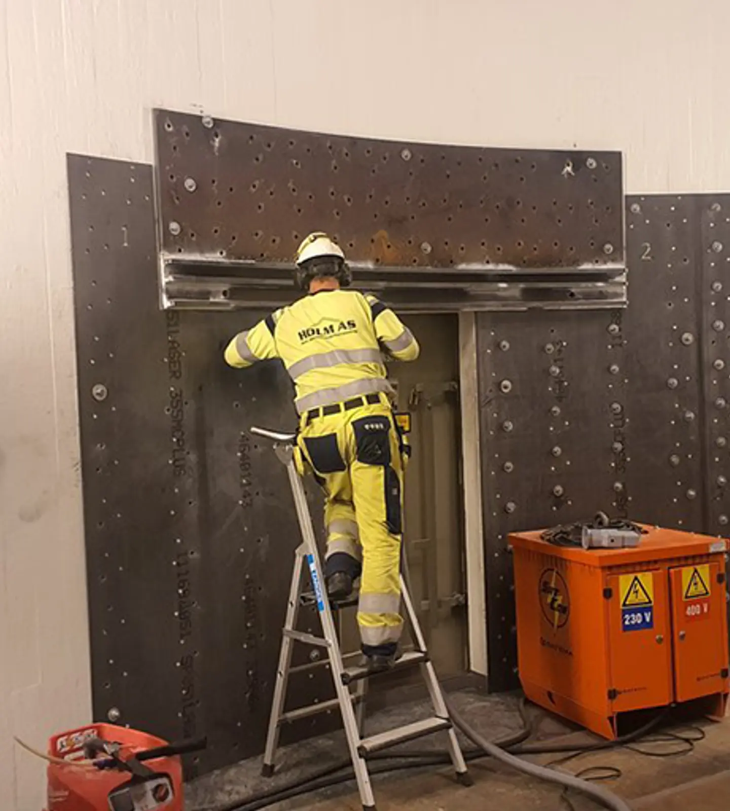 Ansatt i gul vernedress og hjelm jobber på en stige med en stålkonstruksjon inne i et anlegg