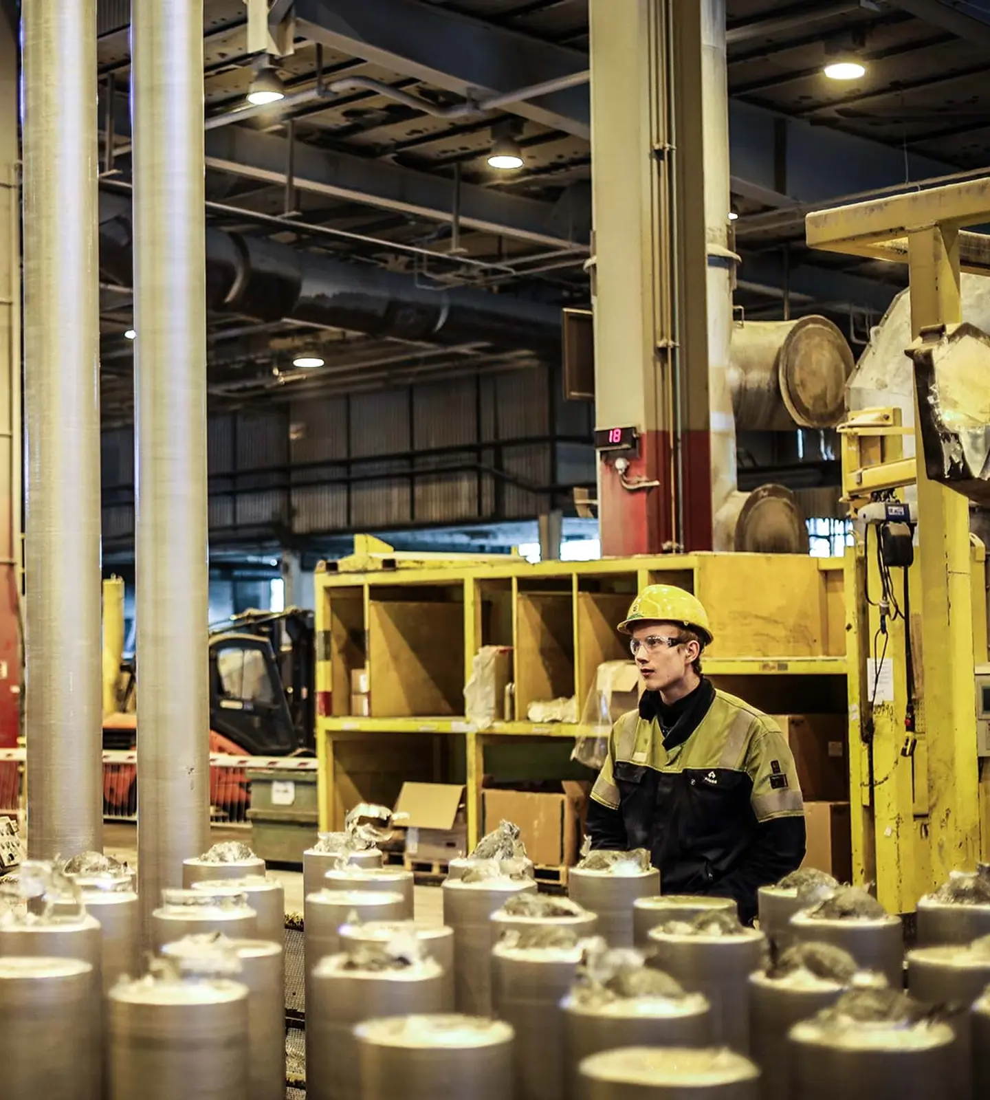 Ansatt med hjelm og vernebriller ved produksjonslinjen på Alcoa Lista, omgitt av aluminiumsstenger