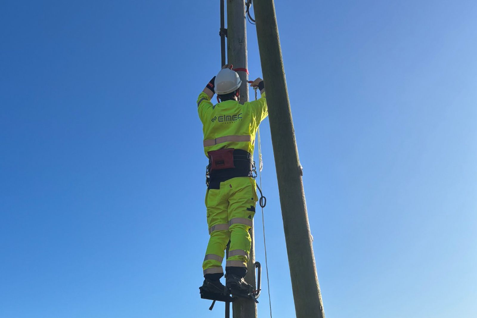 En montør i gul vernedrakt og hjelm klatrer i en høyspentmast mot klar blå himmel.