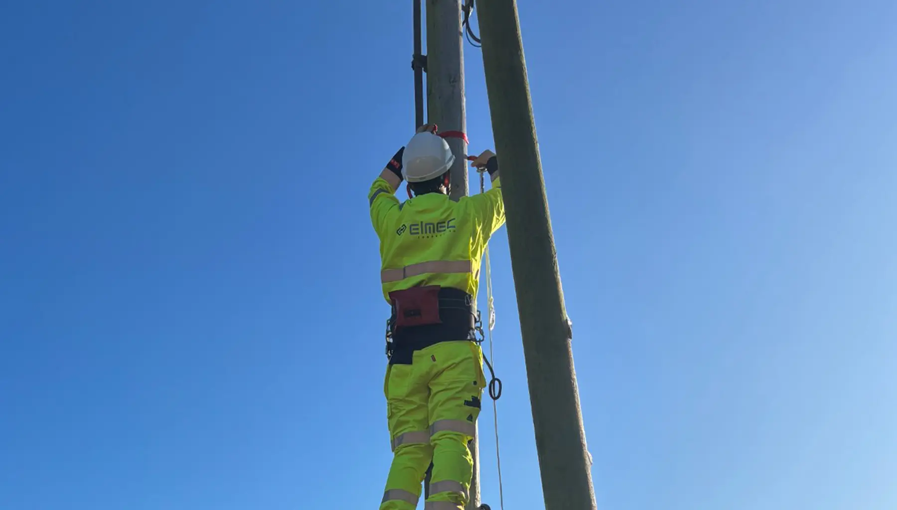 En montør i gul vernedrakt og hjelm klatrer i en høyspentmast mot klar blå himmel.