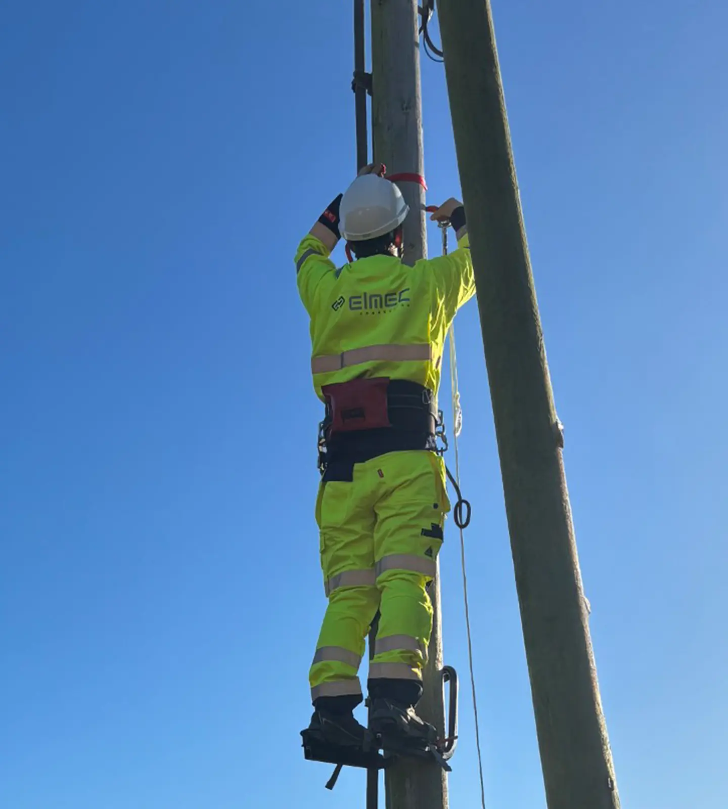 A technician in a yellow protective suit and helmet climbs a high-voltage pylon against a clear blue sky.