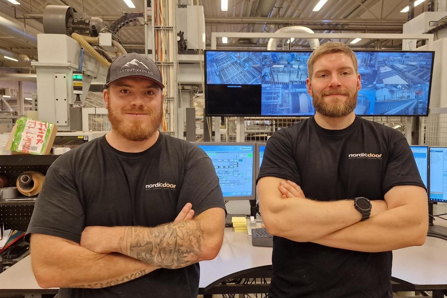 To ansatte i arbeidsklær fra Nordic Door står med armene i kors foran produksjonsutstyr og skjermer i fabrikkhallen