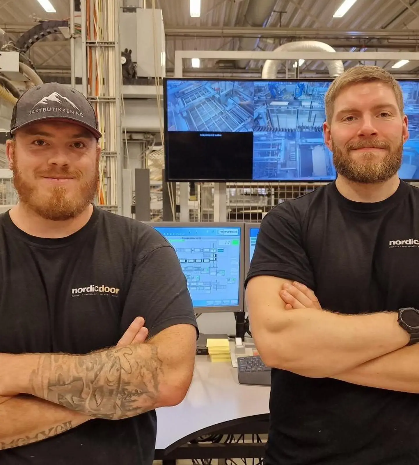 To ansatte i arbeidsklær fra Nordic Door står med armene i kors foran produksjonsutstyr og skjermer i fabrikkhallen