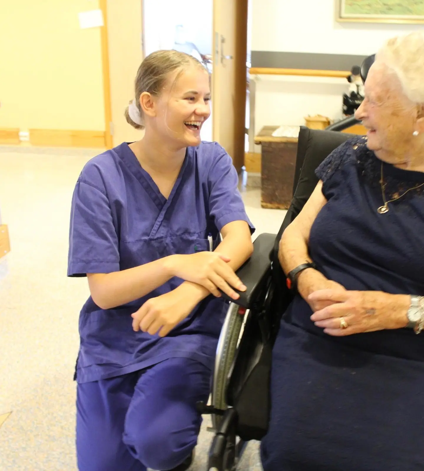 En ansatt i uniform sitter på huk og ler sammen med en eldre dame i rullestol på et sykehjem
