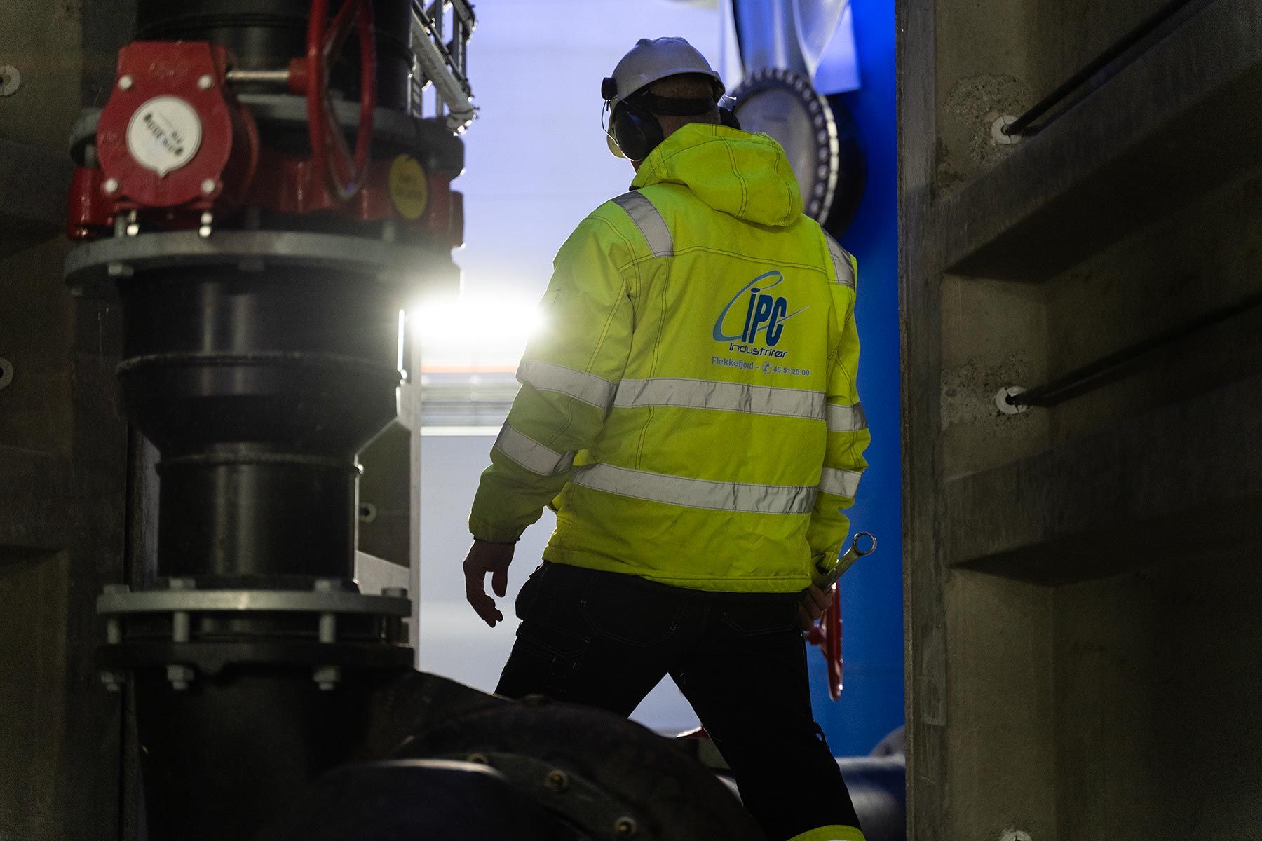 Industrial pipefitter in a yellow safety jacket with the IPC logo walking toward the light inside an industrial facility