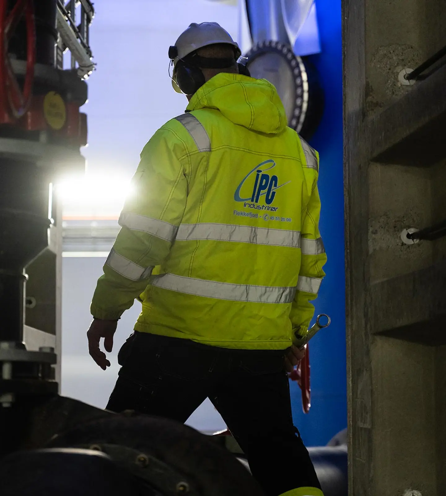 Industrirørlegger i gul vernejakke med IPC-logo går mot lyset inne i et industrianlegg