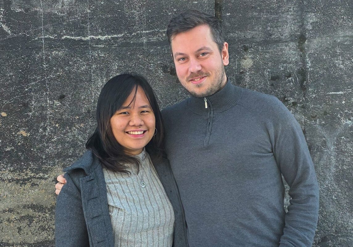 Richard Strømland Egeli og Edlynne smiler mot kamera foran en steinvegg