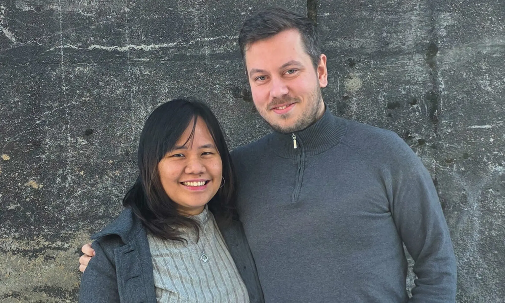 Richard Strømland Egeli og Edlynne smiler mot kamera foran en steinvegg