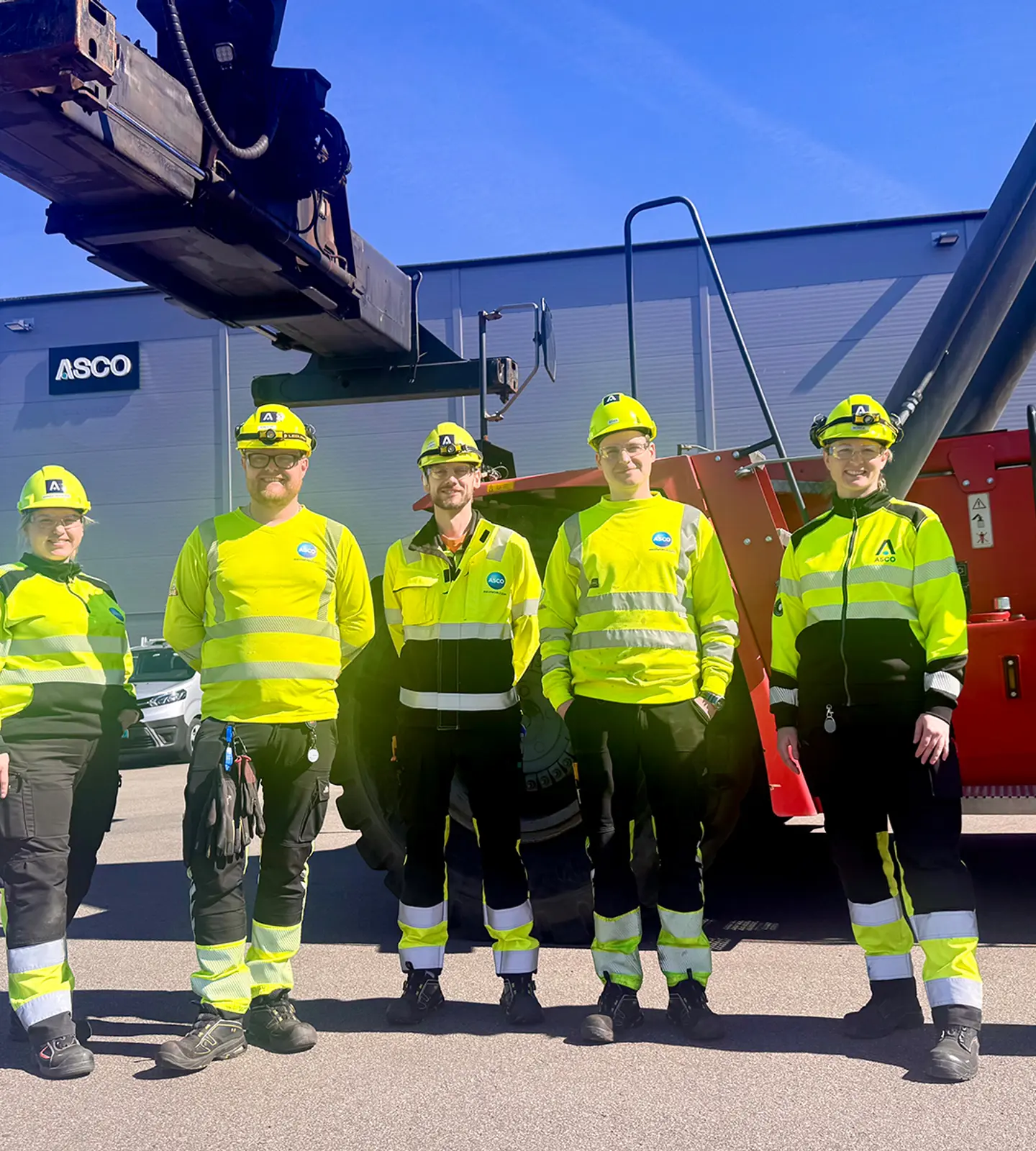 Seks ansatte i gule arbeidsklær og hjelmer står foran en container og en stor gaffeltruck utenfor Asco-anlegget i Farsund.