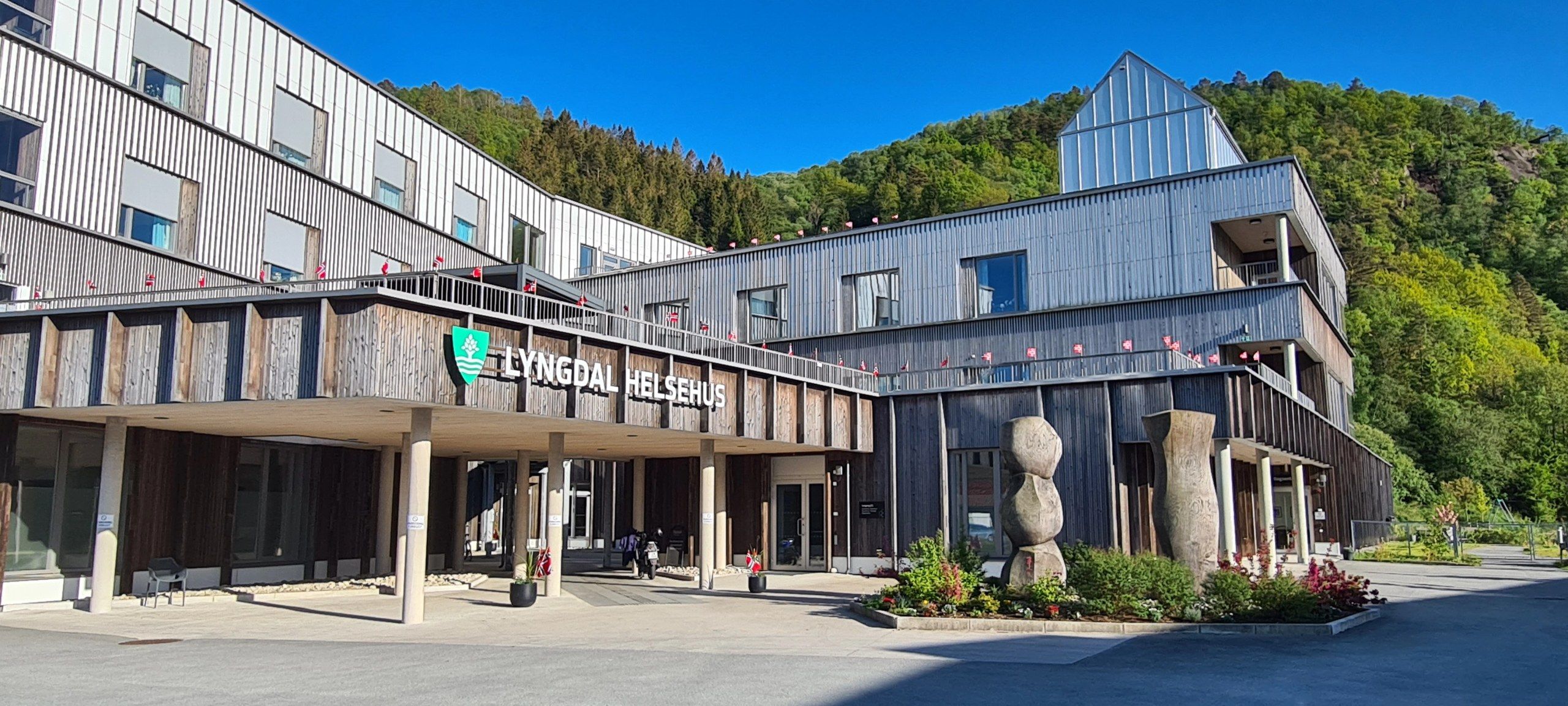 Lyngdal helsehus, et moderne trekledd bygg med inngangsparti og flaggborg, omgitt av skog