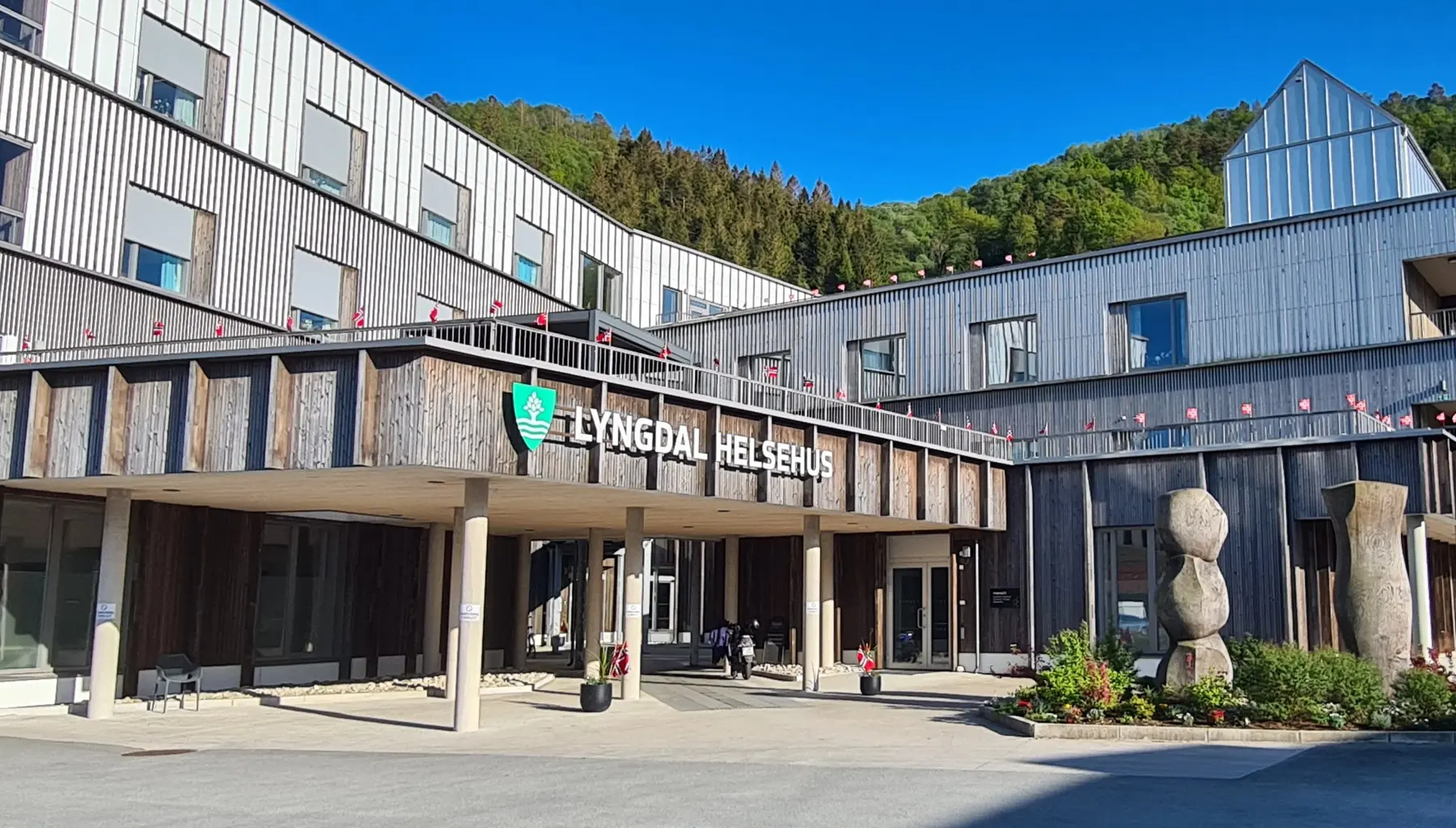 Lyngdal helsehus, et moderne trekledd bygg med inngangsparti og flaggborg, omgitt av skog