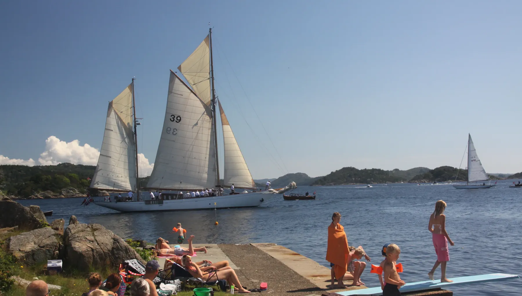 Badende barn og voksne på svabergene ved Badehuset i Farsund, med stupebrett og en seilskute for fulle seil rett utenfor