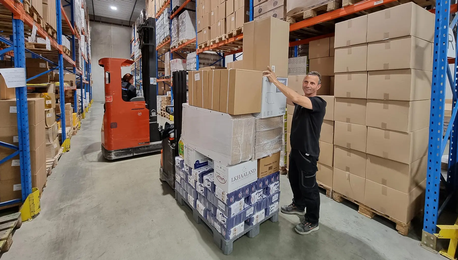 En ansatt henter varer fra en pall i lageret til PB Distribusjon, med en trucksjåfør i bakgrunnen