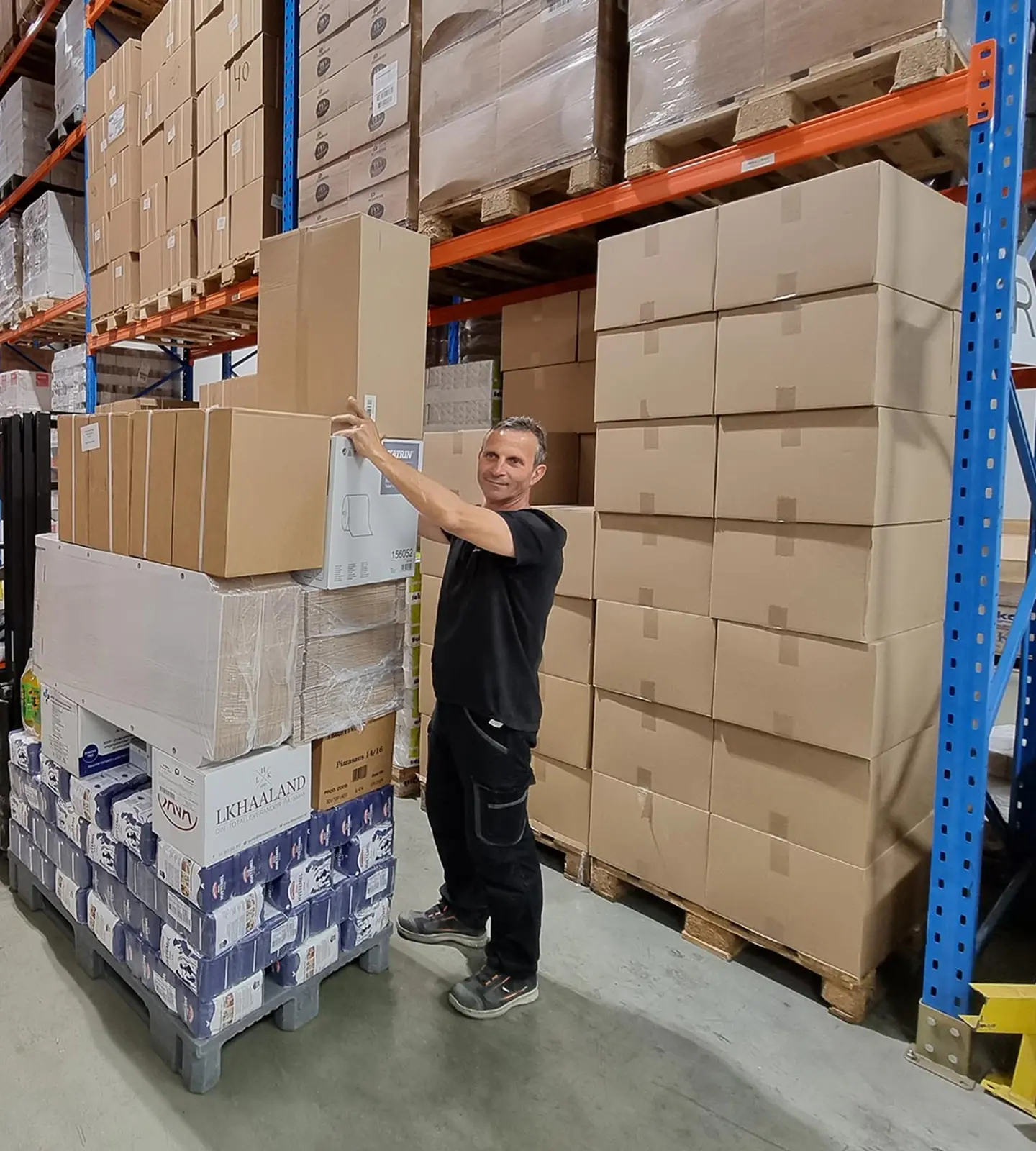 En ansatt henter varer fra en pall i høyreolelageret til PB Distribusjon, med en trucksjåfør i bakgrunnen