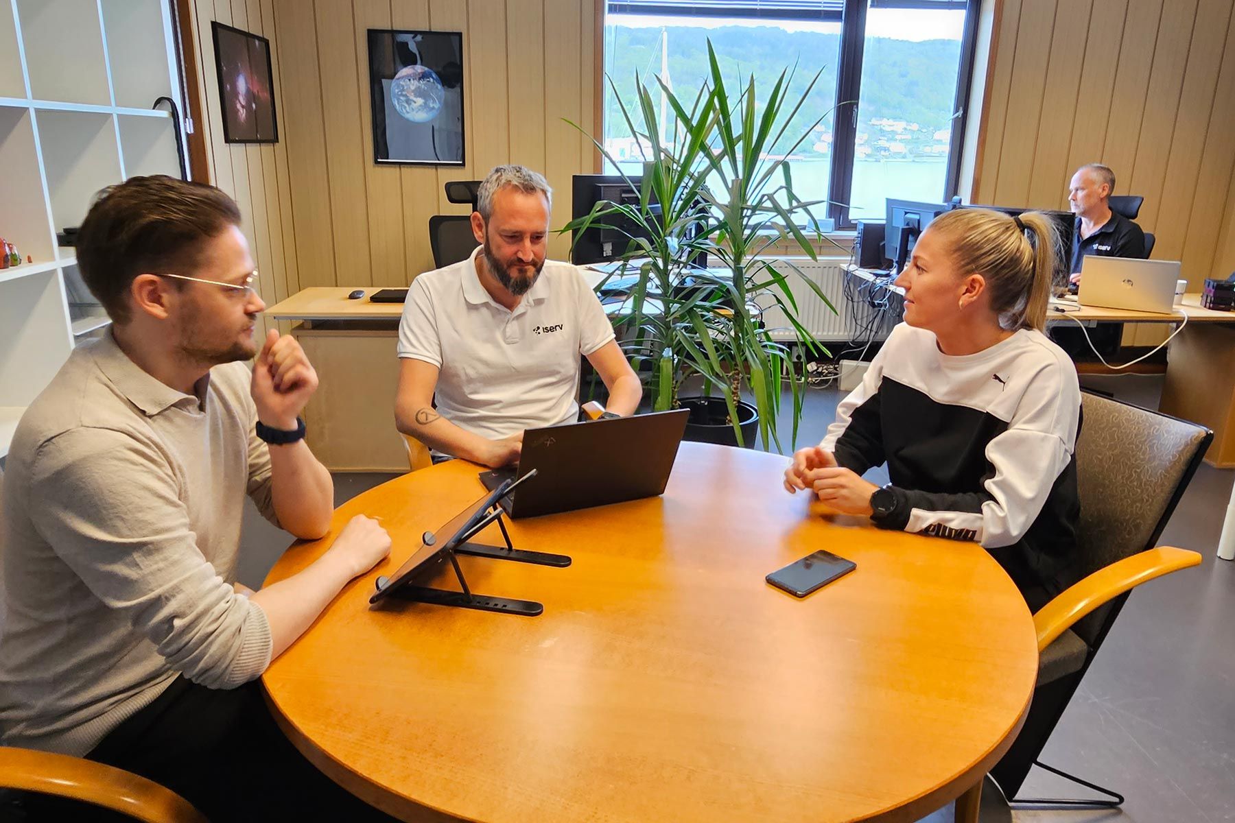 Tre kolleger i samtale rundt et møtebord på Iservs kontor, med utsikt over fjorden i bakgrunnen