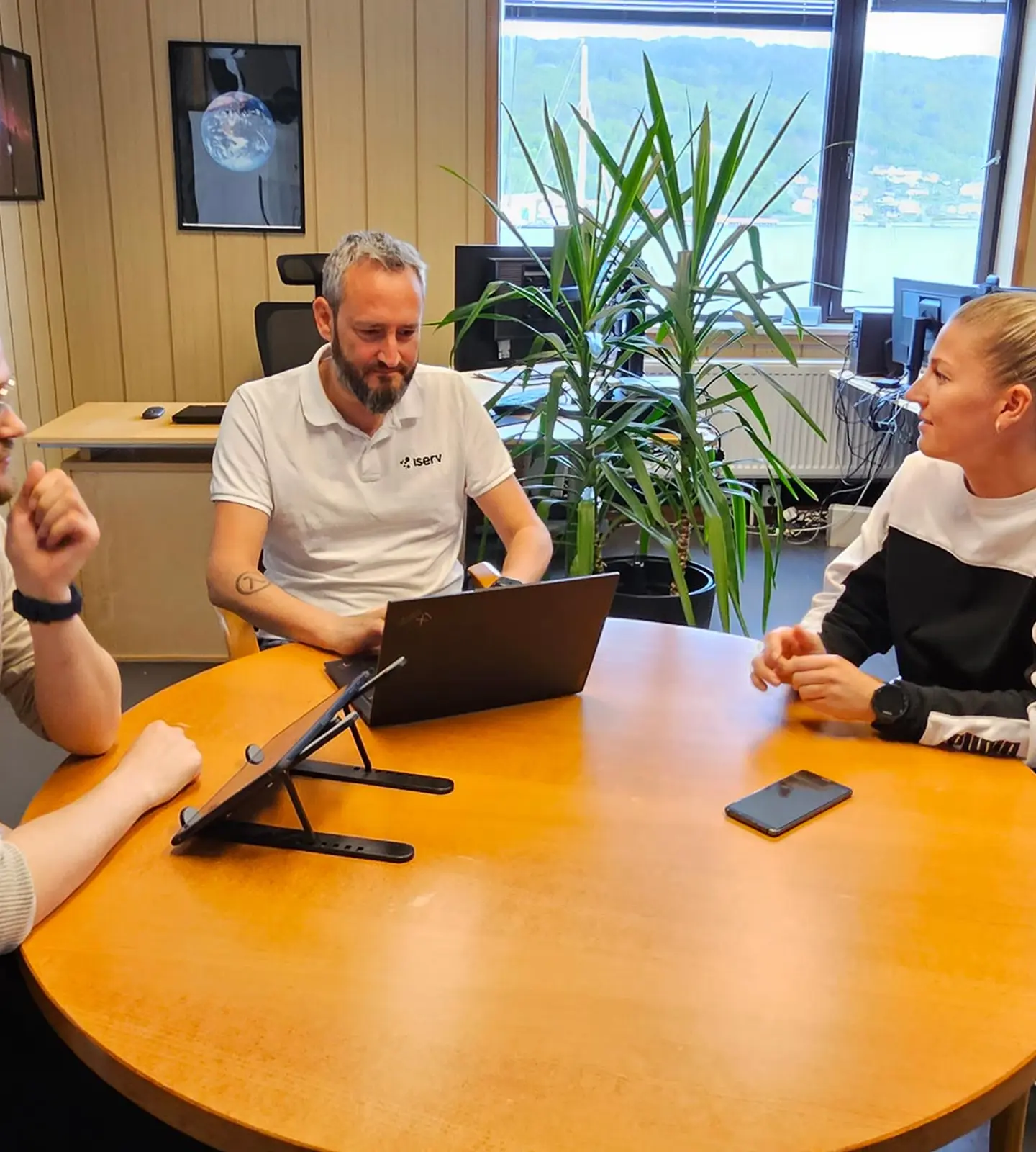 Tre kolleger i samtale rundt et møtebord på Iservs kontor, med utsikt over fjorden i bakgrunnen