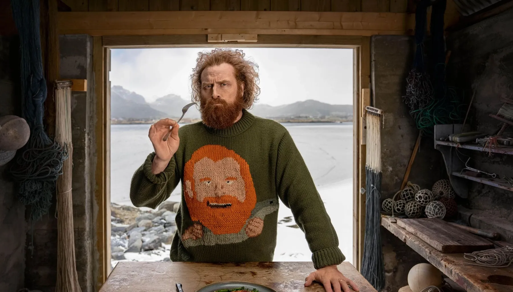 Mann spiser en laksemiddag ved et trebord i en sjøbu med fjord i bakgrunnen