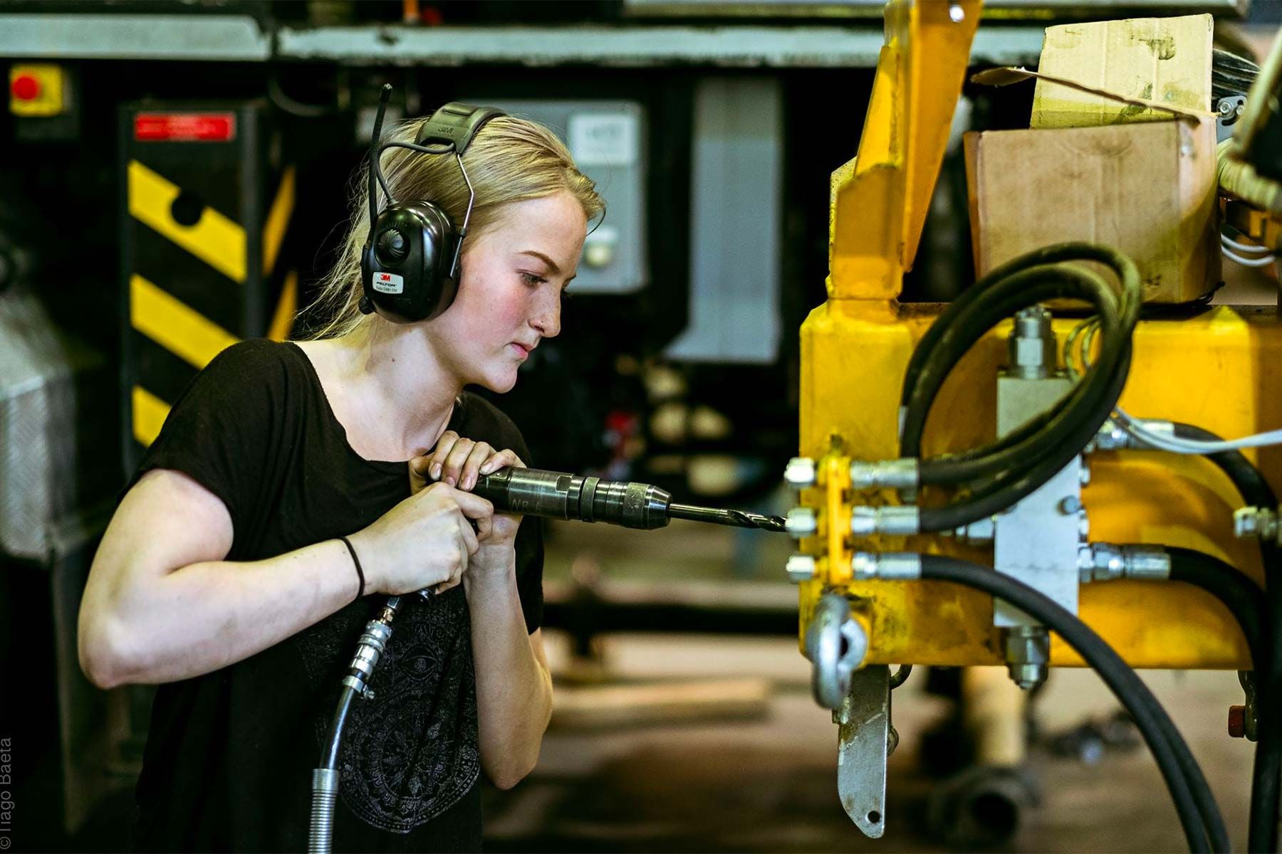 Ung kvinnelig fagarbeider med hørselvern borer i en gul hydraulikkomponent på en maskin i produksjonshallen hos AMV.