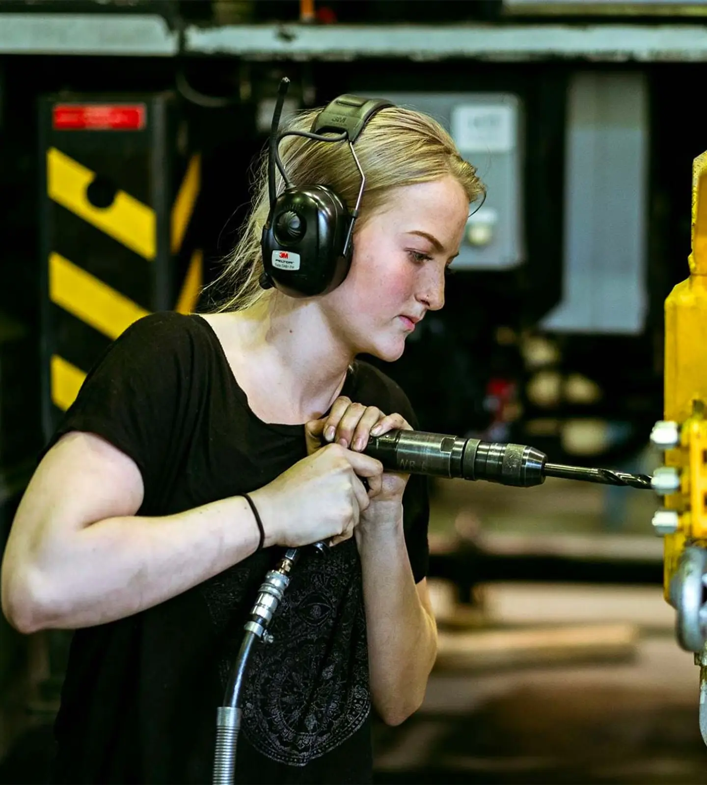Ung kvinnelig fagarbeider med hørselvern borer i en gul hydraulikkomponent på en maskin i produksjonshallen hos AMV.