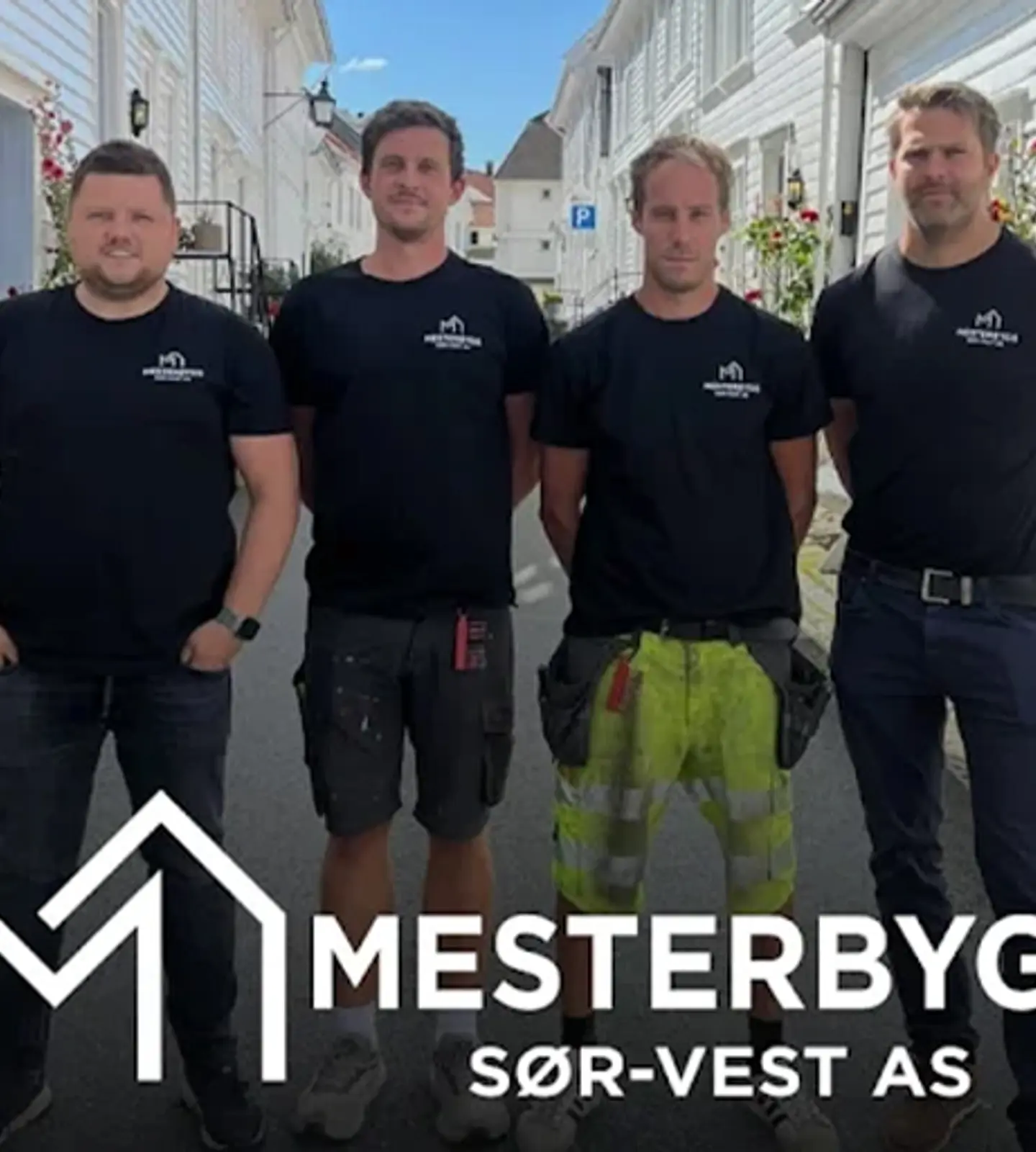 Fire ansatte i Mesterbygg Sør-Vest AS står samlet i en gate med hvite trehus, foran firmaets logo