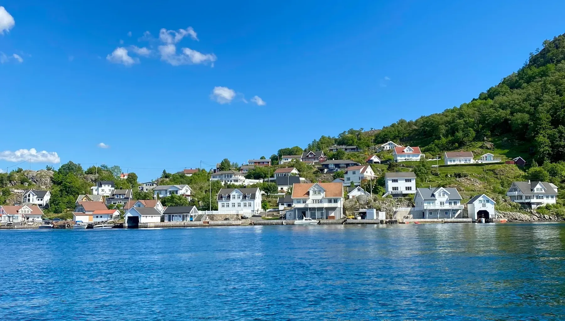 Bygda på Andabeløy sett fra fjorden, med hvite hus under en grønn ås og blå himmel